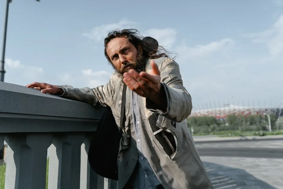 A homeless man in tattered clothing extends his hand, pleading for help on a sunny day.