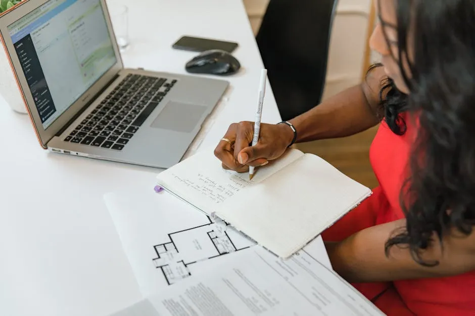 Businesswoman in office writing notes while working on laptop for project planning.